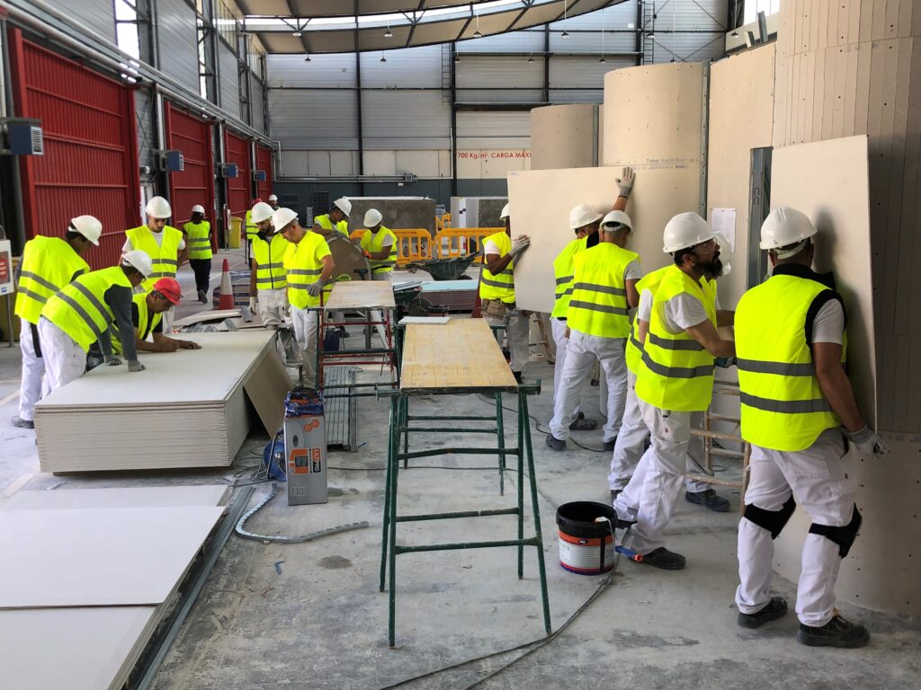 Grupo de alumnos en formación instalando placas de yeso laminado en estructuras curvas dentro de un taller práctico. Todos llevan equipo de protección individual con cascos y chalecos de alta visibilidad.