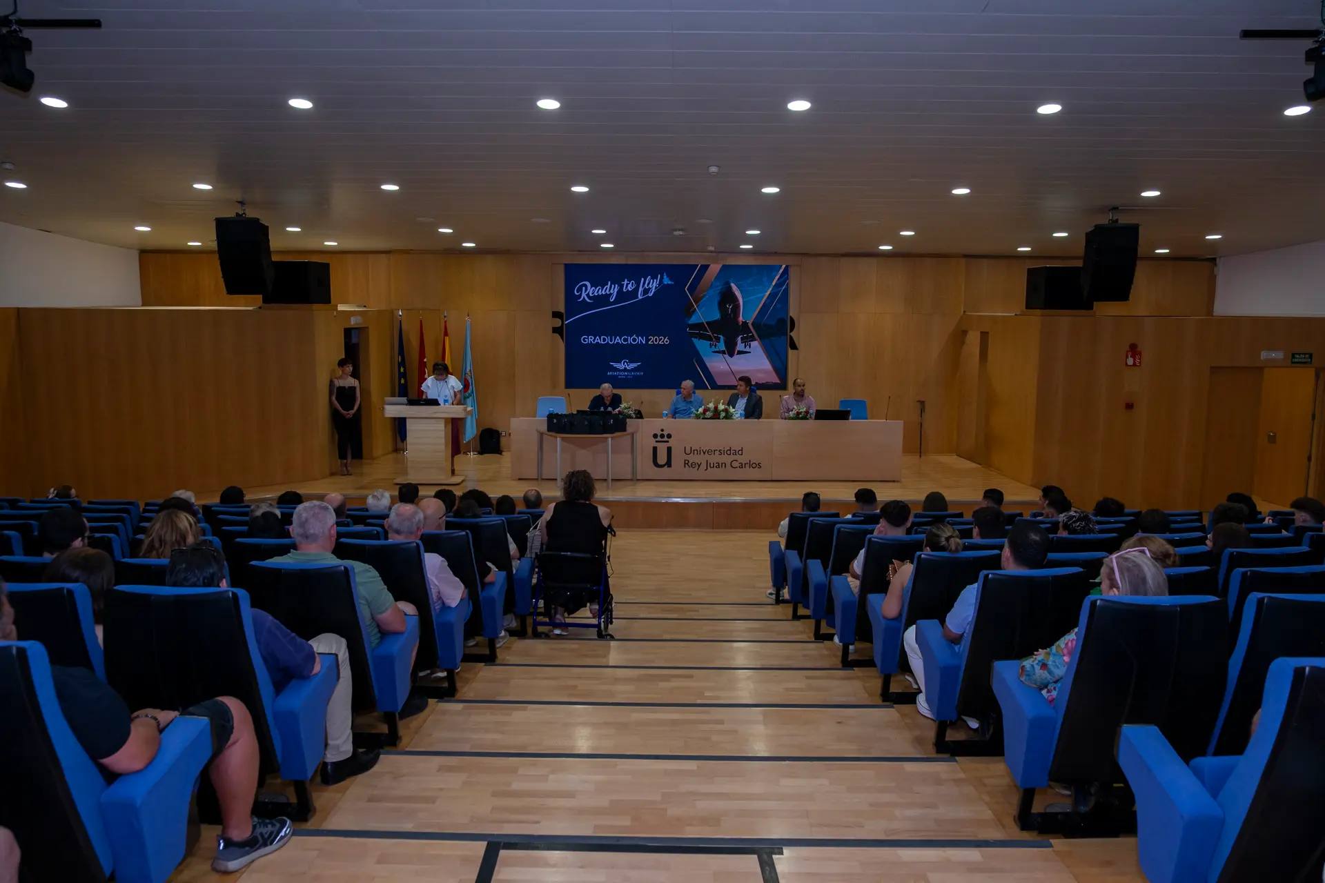 Vista de auditorio universitario con público en butacas azules asistiendo a una solemne ceremonia de graduación.