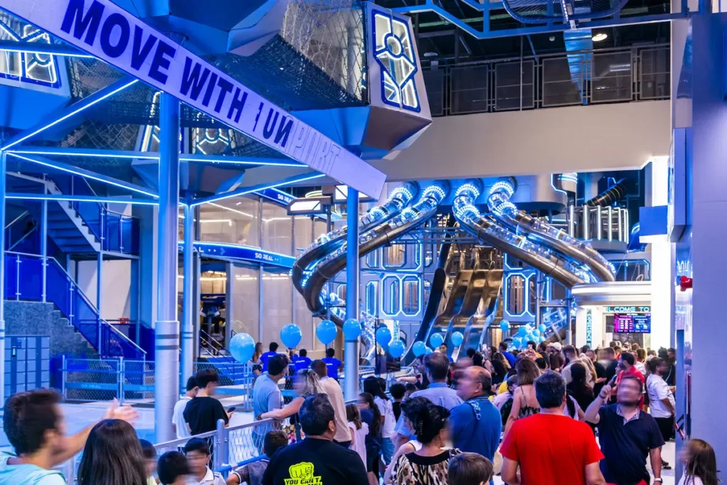 Multitud en centro de ocio interior con diseño futurista, luces azules y grandes toboganes metálicos al fondo.