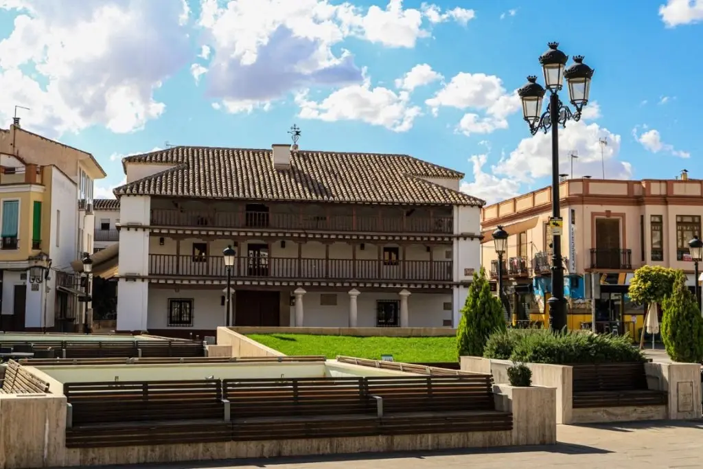 Plaza pública con edificio blanco tradicional de balcones de madera, bancos modernos y farola clásica bajo cielo azul.