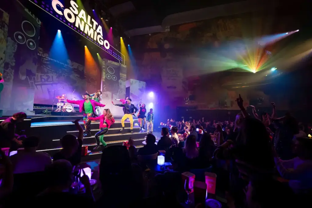 Artistas con ropa colorida retro bailando en el escenario del espectáculo Salta Conmigo frente a un público muy animado.