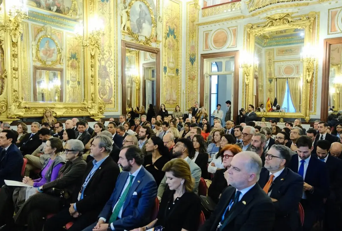 Gran audiencia de asistentes sentados en un evento formal dentro de un salón clásico con decoración dorada y espejos.