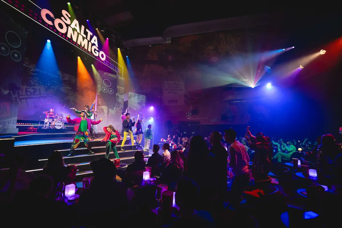 Artistas actúan en el escenario bajo el letrero Salta Conmigo mientras el público disfruta del show desde sus mesas.