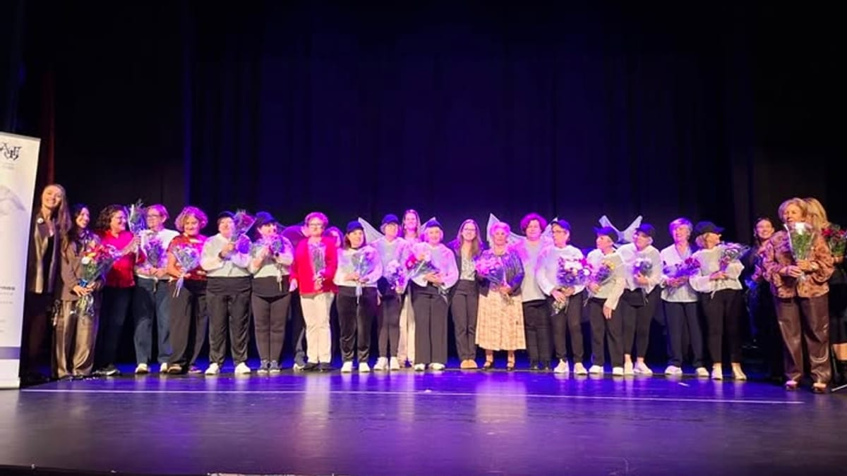 Grupo de personas en un evento de la Asociación de Mujeres Fuenlabreñas sosteniendo flores púrpuras bajo iluminación azul en escenario