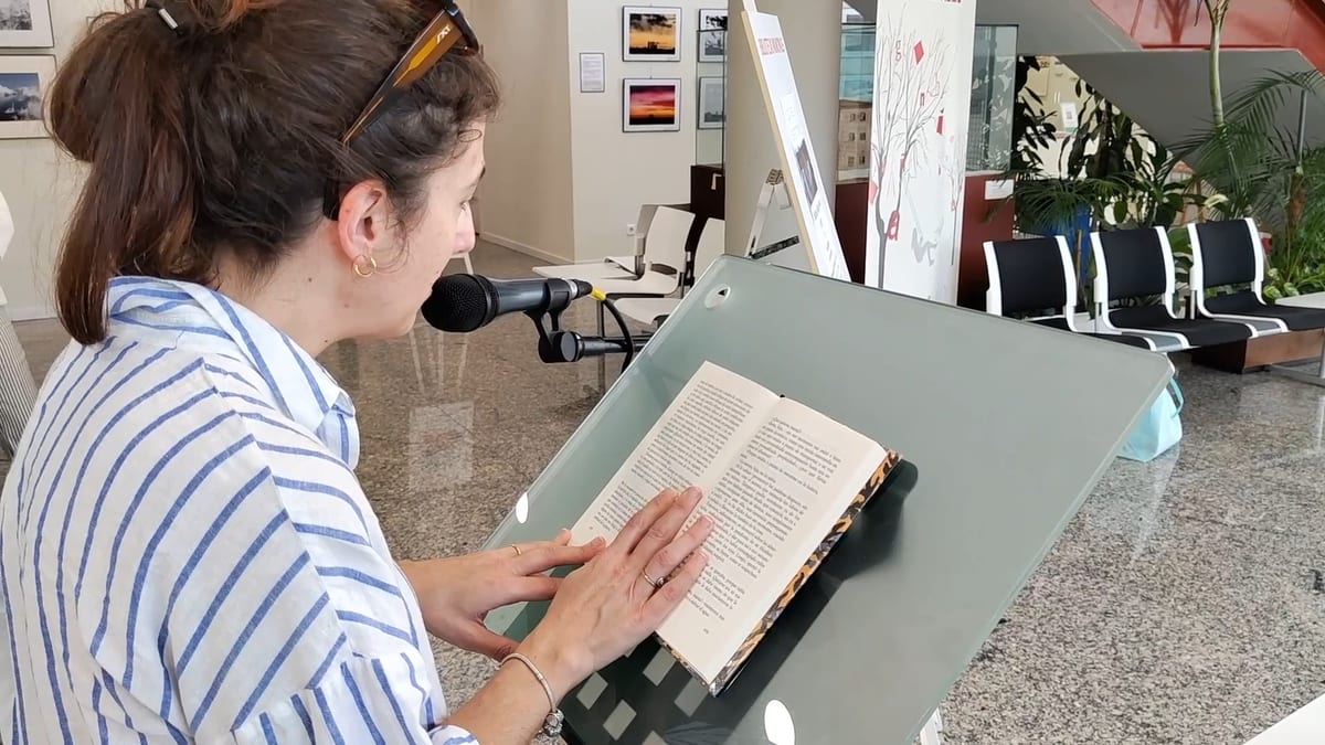 Persona leyendo de un libro mientras sostiene micrófono en acto de lectura pública en la Biblioteca Marcos Ana
