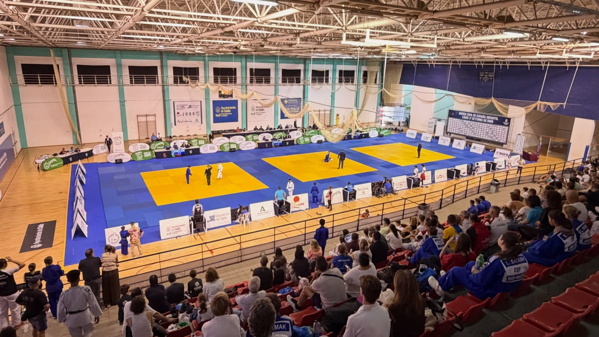 Evento de judo con múltiples tatamis azules y amarillos en un gimnasio cubierto, espectadores en gradas y banners de patrocinadores