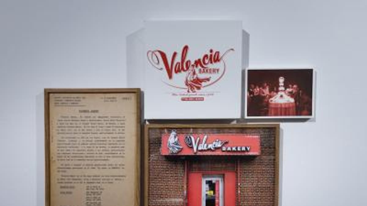 Instalación artística de la exposición 'Un vacío en Nueva York' con elementos visuales relacionados con Valencia, incluyendo un letrero de neón rojo y documentos enmarcados