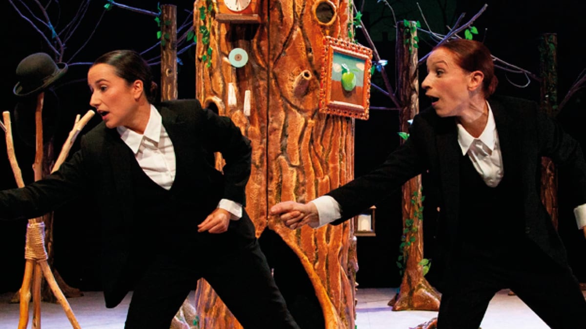 Escena de performance teatral con dos actores en trajes negros ante una estructura decorada de árbol, parte del programa Danza a Escena