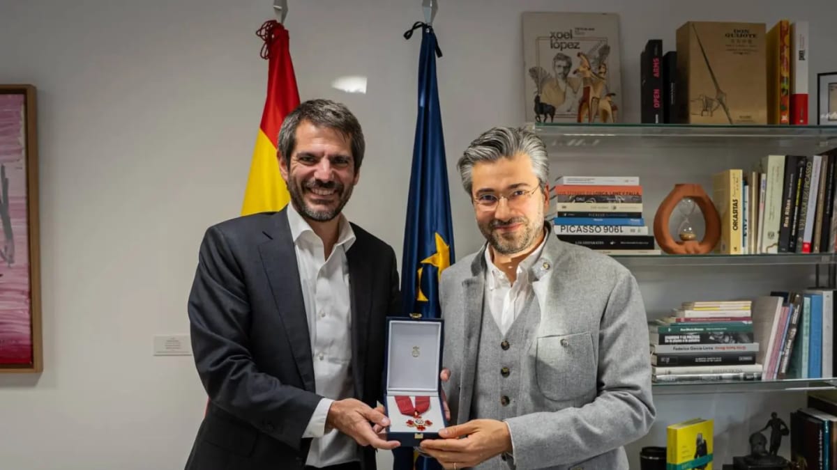 David Afkham sostiene la condecoración de la Encomienda de Alfonso X el Sabio junto a un responsable institucional en una oficina con banderas de España y la UE