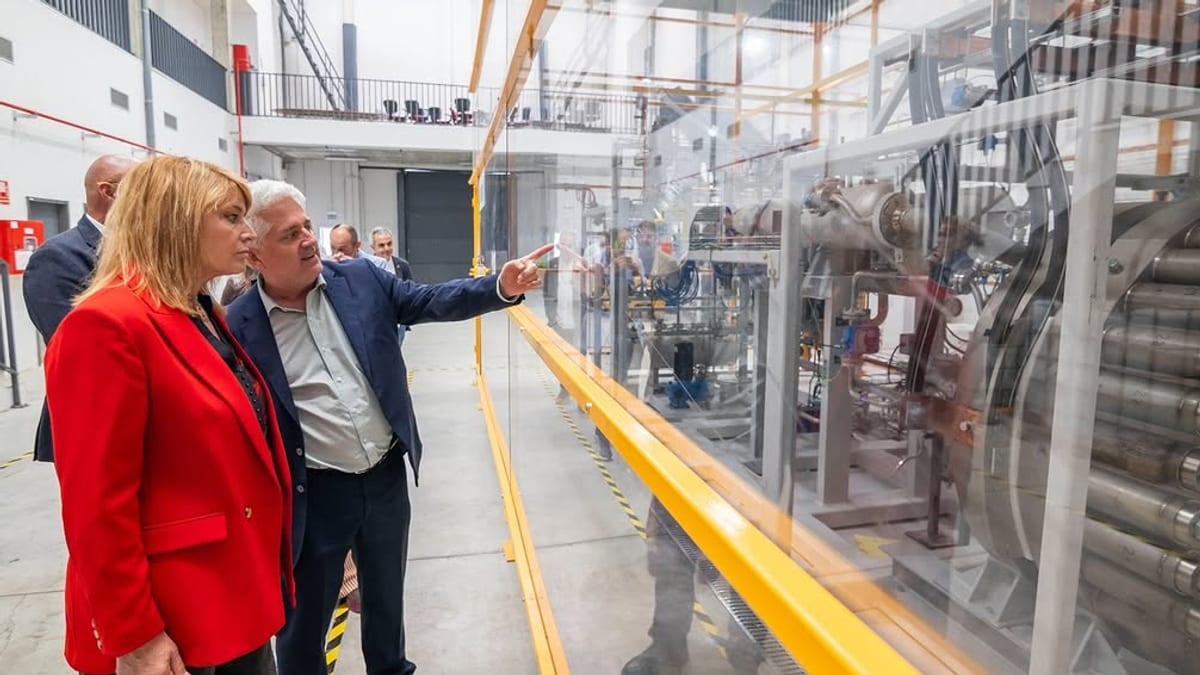 Dos personas observan una instalación industrial con maquinaria a través de cristal, relacionada con tecnología de hidrógeno