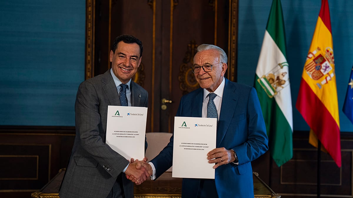 Juanma Moreno e Isidro Fainé realizan un apretón de manos mientras sostienen documentos, con banderas de España y Andalucía de fondo en un acto oficial