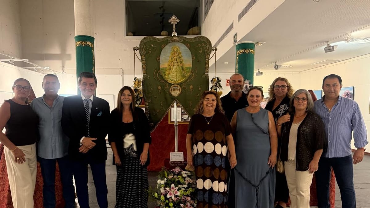 Grupo de personas en la inauguración de la exposición 'El Rocío con el color de los sesenta' frente a estructura religiosa decorada en Punta Umbría