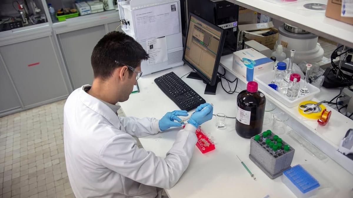 Trabajador de laboratorio en bata blanca utilizando guantes azules, trabajando con muestras en bandejas de laboratorio