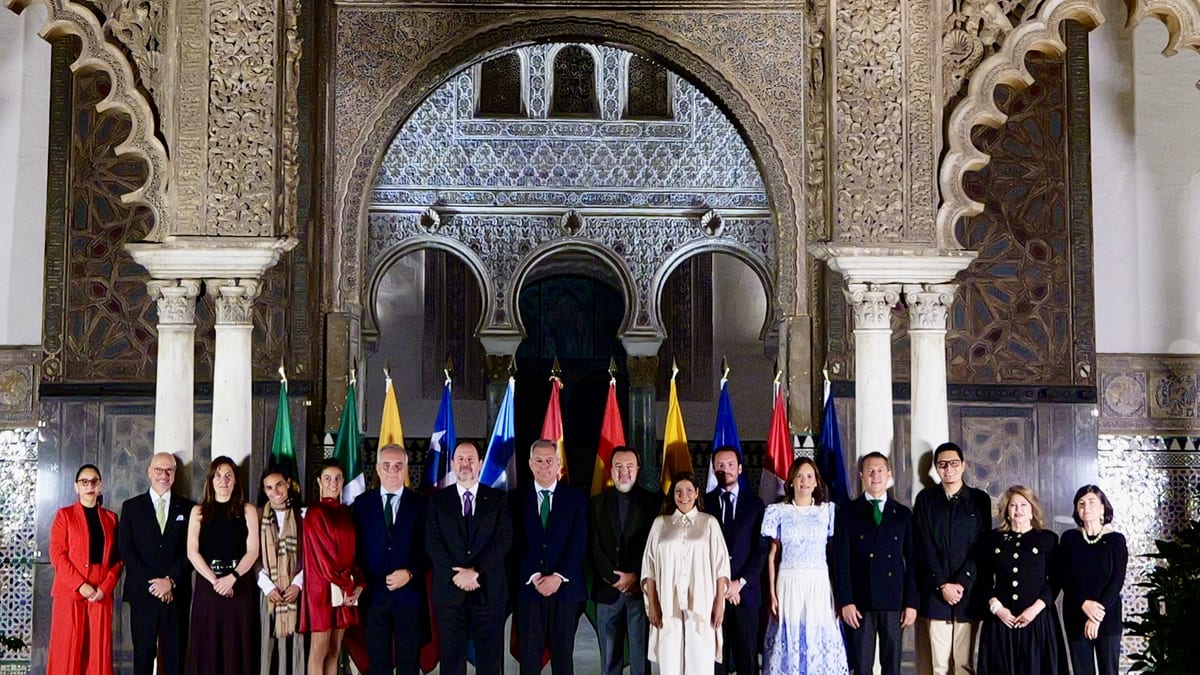 Grupo de alcaldes iberoamericanos posando formalmente en el Real Alcázar de Sevilla con banderas de diferentes países al fondo